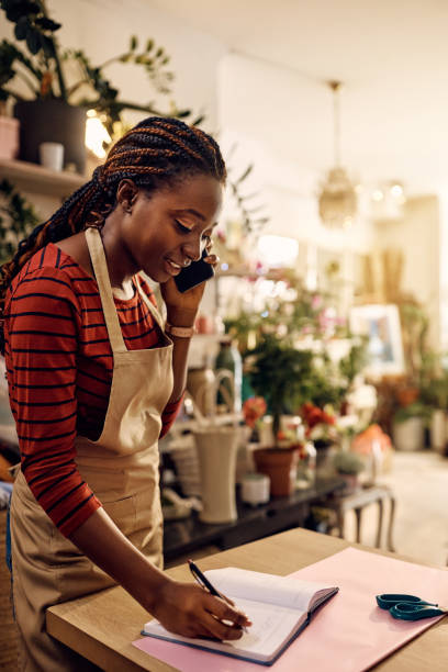 istock Black female florist talking on mobile phone and taking notes while receiving order at flower shop. 1401396350