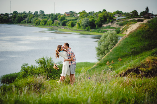 Loving couple kissing against the background of the river and green nature. Love story of young man and woman. Beautiful girl with long curly hair hugging with her boyfriend outdoors