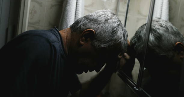 Desperate senior leaning head on bathroom mirror suffering from illness stock photo