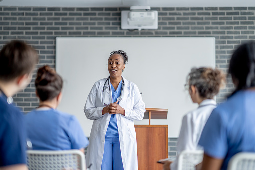 A female doctor of African decent, stands at the front of the room as she teaches a room full of students.  She is wearing blue scrubs and has a lab coat over top as she teaches the class.  The students are dressed professionally in scrubs and seated in front of her as they listen attentively.