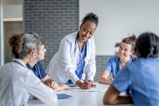 A mature female doctor of African decent, stands at the head of a table as she meets with her team to discuss her patients needs.  She is wearing a white lab coat and the other team members are all wearing scrubs.  They are sitting and listening attentively.