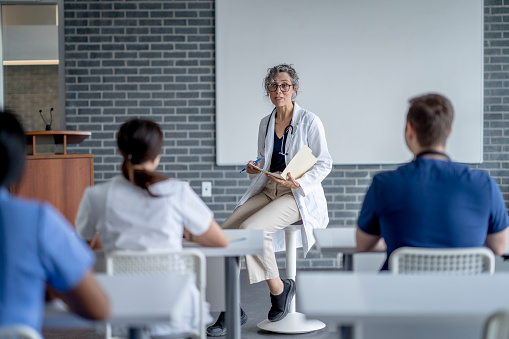 A mature female doctor sits at the front of a classroom as she teaches a small group of students.  She has a file folder on her lap as she talks with the students, and is dressed in a white lab coat.  The students are seated in front of her and paying close attention as they take notes.