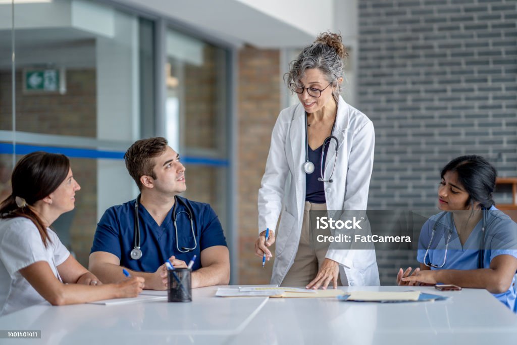 Medical Team Meeting A mature female doctor stands at the head of a table as she meets with her team to discuss her patients needs.  She is wearing a white lab coat and the other team members are all wearing scrubs.  They are sitting and listening attentively. Nurse Stock Photo