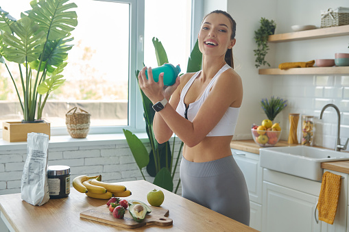 Happy young woman in sports clothing preparing protein cocktail at home