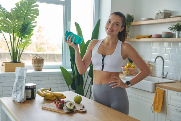 Cheerful young woman in sports clothing preparing protein cocktail at home stock photo