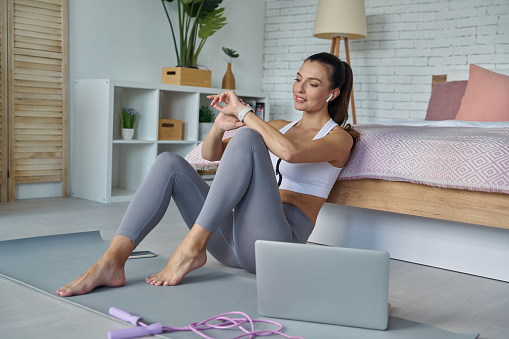 Happy woman in sports clothing looking at smart watch while sitting on the floor at home