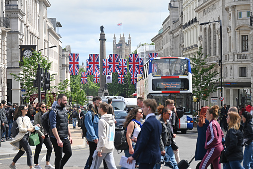 People passing through Piccadilly Circus in London with the backdrop of British flags, distant Houses of Parliament and a tourist bus. London, UK