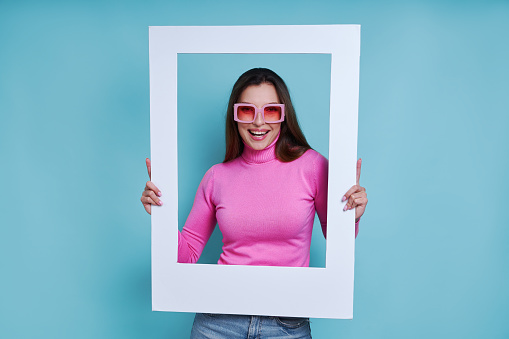 Happy young woman looking through a picture frame while standing against blue background