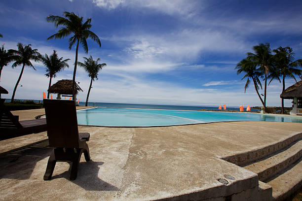 Swimming pool surrounded by concrete deck with two wooden chairs against ocean backdrop with blue sky and clouds with Palm Trees