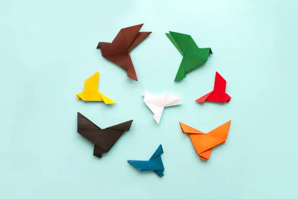 Photo of seven paper origami pigeons different colors in circle fly away from white bird on light blue background