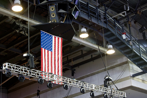 Looking up at sports arena construction, stage truss, lighting components and sound equipment, featuring American Flag.