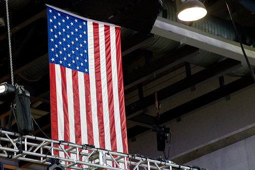 Looking up at sports arena construction, stage truss, lighting components and sound equipment, featuring American Flag.