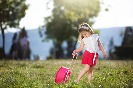Portrait of a happy little blonde girl posing with pink skirt, white blouse and pink suitcase in a publick park