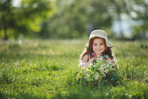 Portrait of a happy little blonde little girl in a colorful floral jumpsuit and a hat holding a basket of flowers, she is sitting on grass