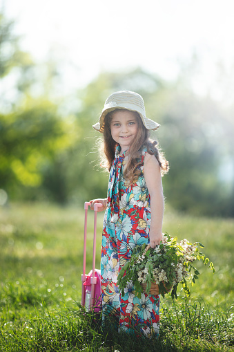 Portrait of a happy little blonde little girl in a colorful floral jumpsuit holding a basket of flowers and suitcase