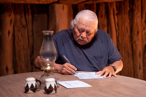 Senior Adult Navajo Man Sitting At The Table In His Hogan Filling Out His Mail-In Ballot For The Upcoming Election