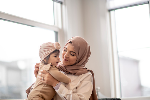A single Muslim Mother lifts her baby up and in close to her. They are both wearing muted neutral tones and have traditional headdress on as they smile and enjoy the bonding time.
