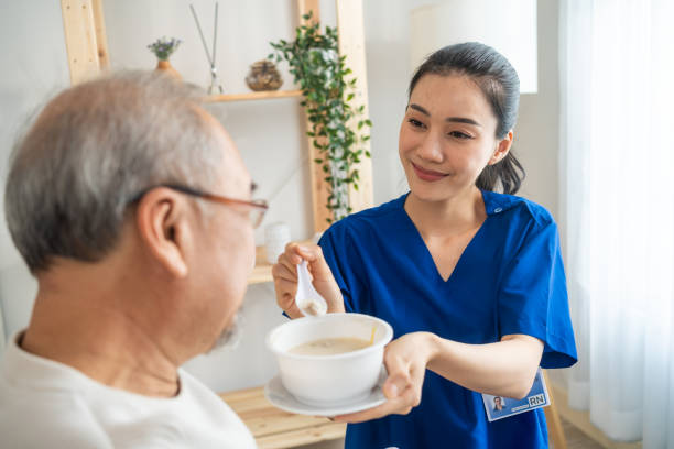 Asian physician nurse support to elderly male patient on wheelchair. Caregiver therapist pharmacist girl serving and feeding food to older patient in living room at home, Healthcare medical concept. stock photo