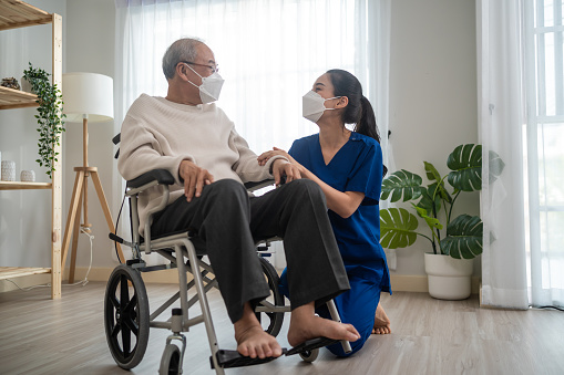 Portrait of Asian caregiver nurse take care senior male on wheelchair. Attractive specialist carer women wear face mask, support elderly grandfather at nursing home care and smile, look at each other.