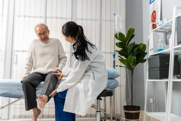 Asian woman physician doing legs raise exercise for senior man patient. Attractive female specialist doctor doing physical therapy procedure for elderly mature male to stretch and recover legs muscle. stock photo