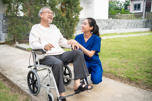 Asian caregiver nurse take care senior male sit on wheelchair outdoor. Beautiful specialist girl doctor help and support elderly mature older patient man doing physical therapy in park at nursing home