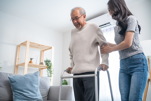 Asian young daughter support senior older man walk with walker at home. Beautiful girl help and take care of elderly mature grandpa patient doing physical therapy for health in living room in house.