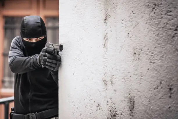 Masked criminal, a fugitive, aiming with a gun, hiding behind a wall