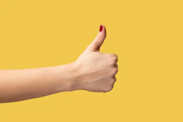 Profile side view closeup of woman hand showing like or thumbs up, approved, positive feedback. Indoor studio shot isolated on yellow background.