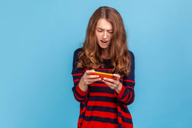 Woman holding smart phone in hands and playing video games with concentrated look, loosing level. Portrait of woman wearing striped casual style sweater, holding smart phone in hands and playing video games with concentrated look, loosing level. Indoor studio shot isolated on blue background. woman defeat stock pictures, royalty-free photos & images