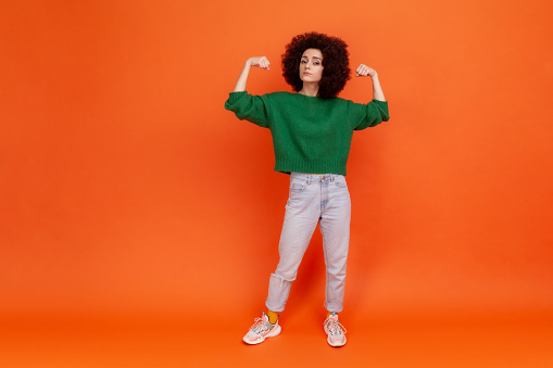 Full length portrait of woman with Afro hairstyle in green sweater raising arms showing biceps as metaphor of female power and independence, feminism. Indoor studio shot isolated on orange background.