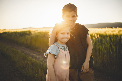 Two kids, brother hugging his little sister on the meadow outdoors in sunset.