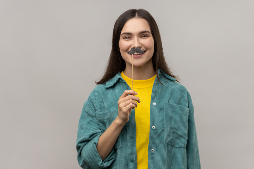Portrait of funny woman with dark hair standing with paper mustache on stick, festive mood, masquerade, wearing casual style jacket. Indoor studio shot isolated on gray background.