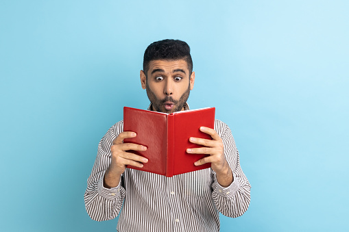 Young adult shocked astonished businessman reading book, reading with big eyes, standing absorbed with amazed plot, wearing striped shirt. Indoor studio shot isolated on blue background.
