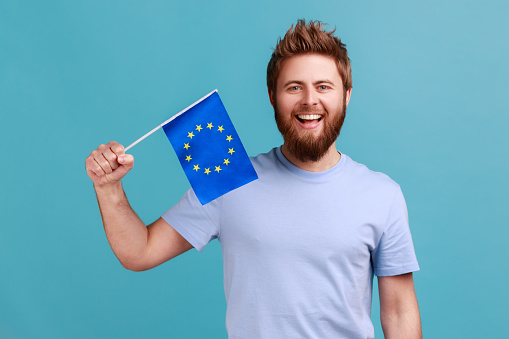 Portrait of excited bearded man smiling broadly and holding flag of European Union, symbol of Europe, EU association and community. Indoor studio shot isolated on blue background.