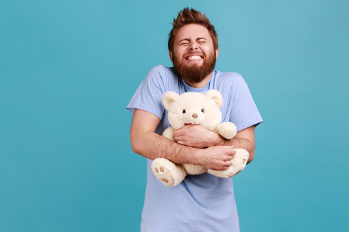 Portrait of satisfied handsome bearded man standing with closed eyes and toothy smile, embracing white soft teddy bear, expressing positive emotions. Indoor studio shot isolated on blue background.