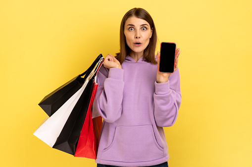 Amazed surprised woman standing with excited look showing shopping bags and smart phone with blank screen for advertisement, wearing purple hoodie. Indoor studio shot isolated on yellow background.