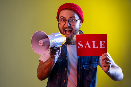 Bearded hipster man paying attention announcing big sale yelling at loudspeaker, black Friday shopping, wearing beanie hat and denim vest. Indoor studio shot isolated on colorful neon light background