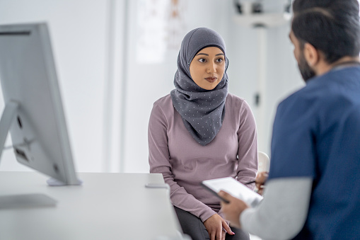 A Muslim woman sits across from her male doctor as she talks with him about her mental health. She is dressed casually and is wearing a Hijab as she sits with a neutral expression on her face. The doctor is wearing blue scrubs and is taking notes on his tablet as the two talk.