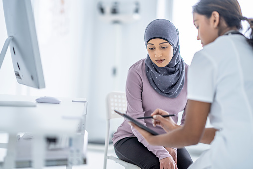 A female doctor sits with a Muslim patient as she reviews her recent test results on a tablet with her. She is seated in white scrubs as she explains the results on the tablet screen to the patient. The patient is seated on a chair in front of her and leaning in to look closely.