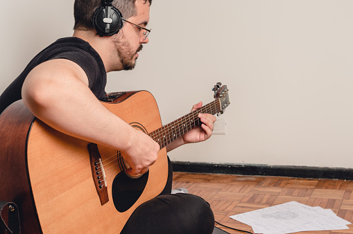 side view young caucasian man with headphones, in online class, studying and playing guitar, reading sheet music sitting on the floor, with the wall in the background and copy space.