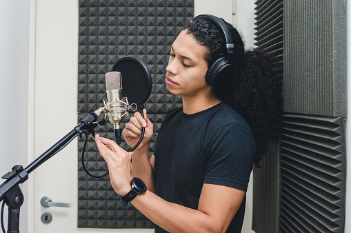 young singer man with headphones inside the professional studio in the recording booth, fixing the anti pop filter and the microphone to sing and record a song.