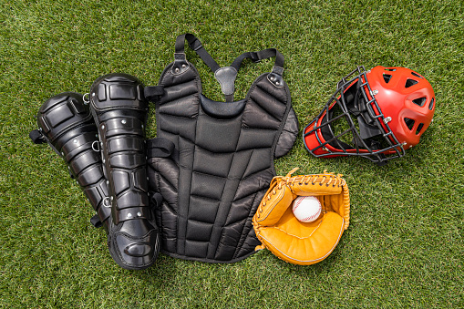 A high angle view of a baseball catcher’s gear consisting of pair of black shin guards, a black chest protector, a red catcher’s mask helmet and a catcher’s mitt with a baseball sitting on the turf.