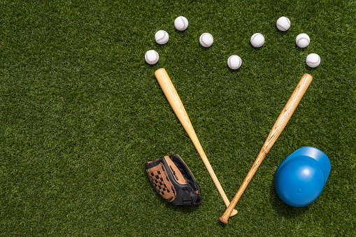 Looking down on new baseballs and 2 wooden bats to form a heart shape, along with a leather glove and a blue batter’s helmet on the artificial turf, for the love of the game