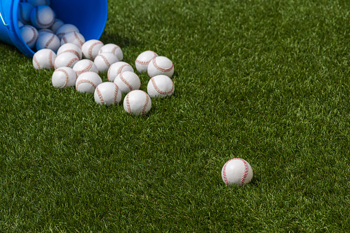 Selective focus on a new baseball that has rolled out of a blue bucket of balls that has spilled on the turf of a baseball diamond, ready for practice.