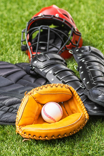 A close-up of a baseball in a catcher’s mitt with the rest of the catcher’s gear sitting on the turf in the background.