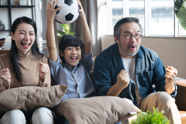 Asian family watching football sports games on TV and reacting happy exiting when team Shoot the ball into the goal. stock photo