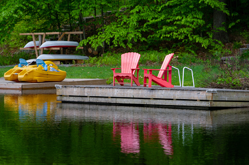 Two red Adirondack chairs on a cottage wooden dock reflecting on the waters of a calm lake in Muskoka.