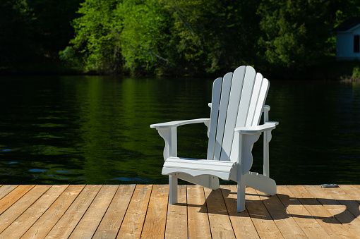 Single white Adirondack chair sitting on a wooden dock in Muskoka Ontario with a calm lake in background.