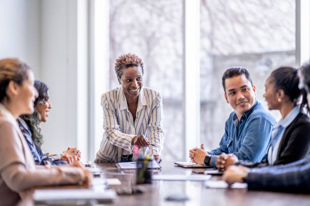 Brainstorming in a Business Meeting A small group of business professionals sit around a table as they meet to brainstorm some ideas for the future of the company. They are each dressed professionally and have papers scattered out in front of them as they work together. The focus is on a woman of African decent who is pitching an idea to the group. meeting stock pictures, royalty-free photos & images