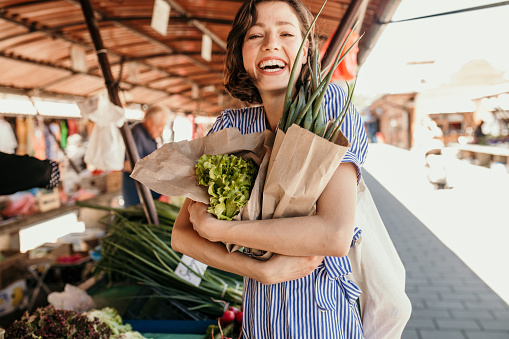 Beautiful woman buying green veggies at a farmer's market. Beautiful smiling female holing a lot of fresh organic vegetables while buying them on a street market.